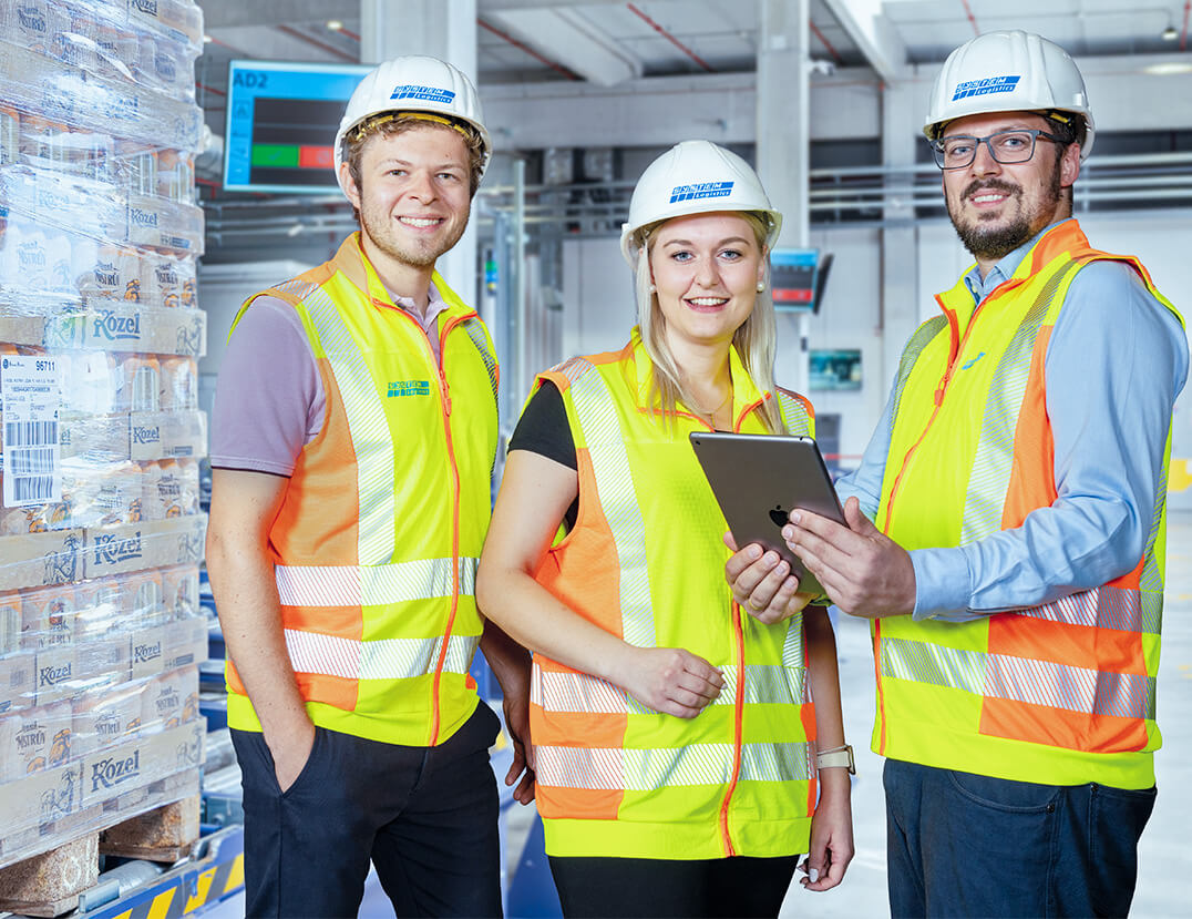 Drei Mitarbeiter von System Logistics mit Warnwesten und Sicherheitshelmen vor beladenen Paletten, ein Mitarbeiter h&auml;lt ein iPad in der Hand