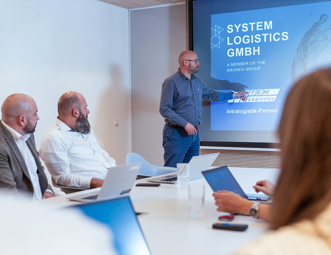 Mitarbeiter von System Logistics pr&auml;sentiert Intralogistikl&ouml;sungen und deutet auf die Leinwand